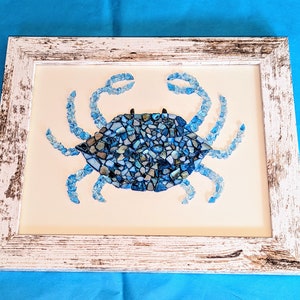 May include: A blue crab mosaic artwork framed in white distressed wood. The crab is made of small pieces of blue and white glass.