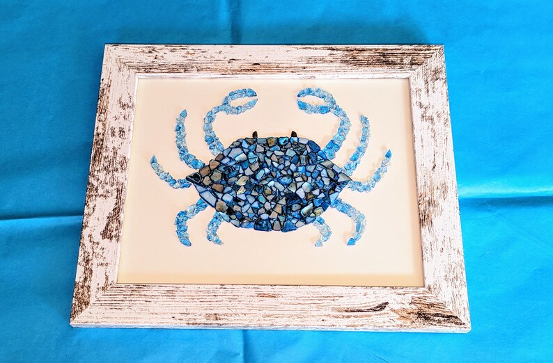 May include: A blue crab mosaic artwork framed in white distressed wood. The crab is made of small pieces of blue and white glass.