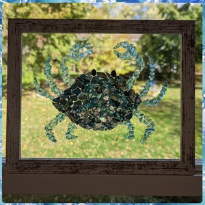 May include: A framed mosaic art piece depicting a crab made from blue and green glass tiles. The crab is set against a blurred background of green foliage.