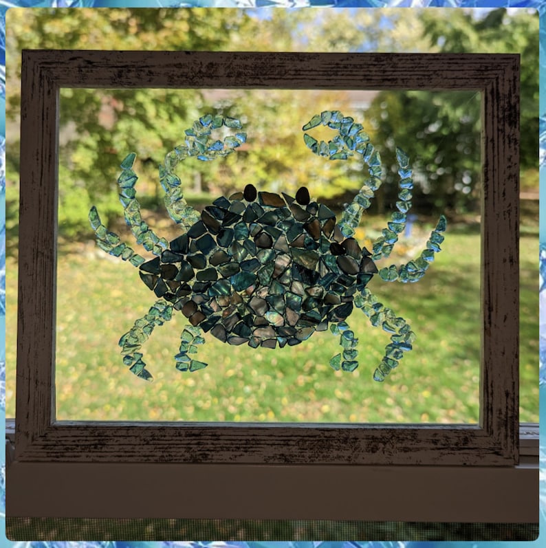May include: A framed mosaic art piece depicting a crab made from blue and green glass tiles. The crab is set against a blurred background of green foliage.