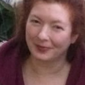 Puede incluir: Una mujer con el pelo rojo sonríe a la cámara. Lleva un suéter morado y una camisa floral.