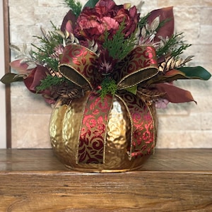 May include: A gold-colored, pumpkin-shaped decorative vase filled with burgundy and green faux flowers and foliage. A red and gold patterned ribbon is tied around the vase. The arrangement sits on a wooden surface.