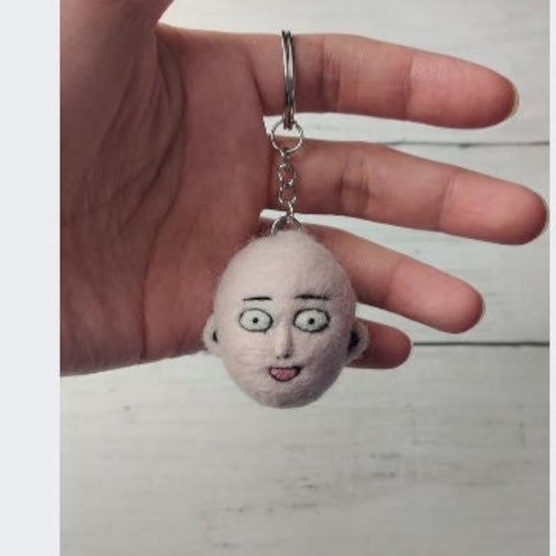 Pode incluir: Um chaveiro de feltro de um homem careca com uma express&atilde;o de surpresa, olhos grandes e a boca aberta. O chaveiro est&aacute; preso a um anel de metal prateado.