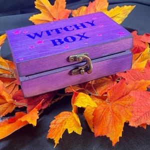 Peut inclure: Une boîte en bois violette avec les mots "WITCHY BOX" en bleu et des étoiles roses. La boîte a un fermoir en métal et est entourée de feuilles d'automne orange et jaune.