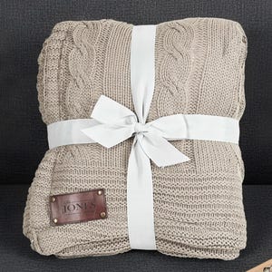 May include: A folded beige cable-knit blanket tied with a white ribbon and bow. A small brown leather tag with the word "JONES" is attached. The blanket is on a dark gray surface.