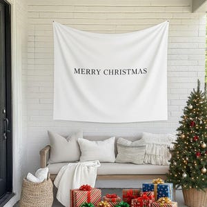 Może przedstawiać: Biały baner z napisem "MERRY CHRISTMAS" wisi nad ławką z poduszkami i kocem. Udekorowana choinka i zapakowane prezenty znajdują się przed ławką, tworząc świąteczną scenę.