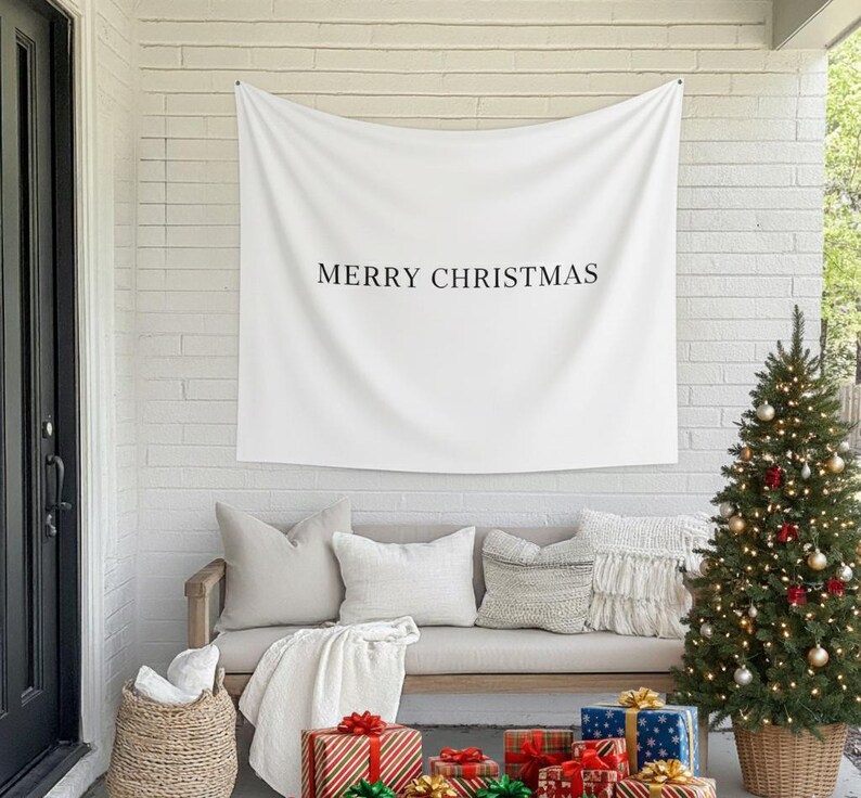 Może przedstawiać: Biały baner z napisem "MERRY CHRISTMAS" wisi nad ławką z poduszkami i kocem. Udekorowana choinka i zapakowane prezenty znajdują się przed ławką, tworząc świąteczną scenę.