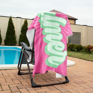 May include: A pink and white striped beach towel with the word "Summer" in large, light green, stylized lettering. The towel is draped over a black folding chair near a swimming pool. The background includes a brick patio and green trees.