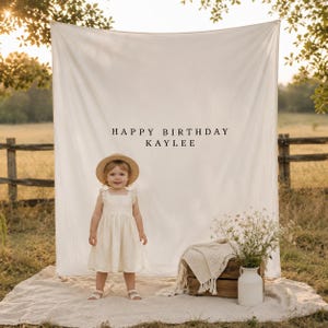 May include: A large white fabric backdrop featuring the black text HAPPY BIRTHDAY KAYLEE, displayed behind a child wearing a cream-coloured dress and a straw hat, with a wooden crate, a white blanket, and a vase of wildflowers nearby.