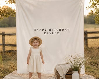 May include: A large white fabric backdrop featuring the black text HAPPY BIRTHDAY KAYLEE, displayed behind a child wearing a cream-coloured dress and a straw hat, with a wooden crate, a white blanket, and a vase of wildflowers nearby.