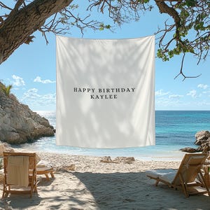 Puede incluir: Una pancarta rectangular blanca con el texto "HAPPY BIRTHDAY KAYLEE" en letras negras. La pancarta est&aacute; colgada en una playa con un cielo azul y el oc&eacute;ano de fondo. Se ven dos tumbonas.