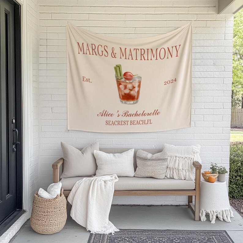 Op de afbeelding: Een witte canvas wandtapijt met de tekst "MARGS & MATRIMONY Est. 2024 Alice's Bachelorette Seacrest Beach, FL" en een tekening van een cocktailglas met een kers en een stengel bleekselderij. Het wandtapijt hangt aan een witte bakstenen muur boven een lichtbruine bank met witte kussens en een wit deken.