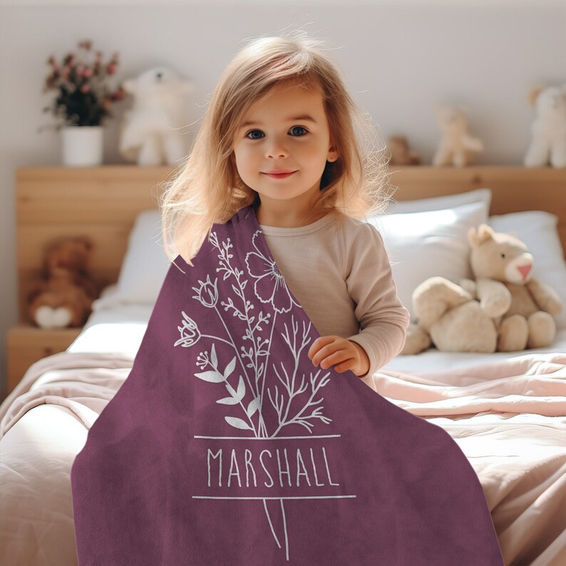 Peut inclure: Une couverture mauve avec un motif floral blanc et le nom "MARSHALL" imprim&eacute; en blanc. Un jeune enfant tient la couverture.