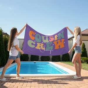 May include: A purple beach towel held by two people, with the words "COUSIN CREW" in colorful, retro-style lettering. The towel also features the name "Cheryl" in cursive. The scene is outdoors near a pool on a sunny day.