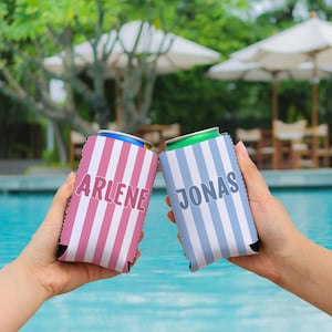 Op de afbeelding: Twee gepersonaliseerde blikjeskoelers die voor een zwembad worden gehouden. De ene koeler is roze met witte strepen en de naam "ARLENE" in wit. De andere is blauw met witte strepen en de naam "JONAS" in wit.