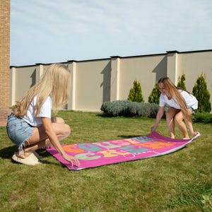 May include: A pink rectangular beach towel with the words "Just Journey" in colorful, retro-style lettering. Two people are unfolding the towel on a green lawn under a sunny sky. The towel is made of a soft material.