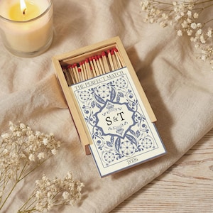 May include: A wooden matchbox with a blue and white floral design. The box contains matches with red tips. The card reads "THE PERFECT MATCH" with the initials "S & T" and the year "2026". A lit candle and white flowers are in the background.