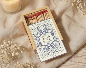 May include: A wooden matchbox with a decorative blue and white floral design. The box contains red-tipped matches. The card reads "THE PERFECT MATCH" with the initials "S & T" and the year "2026". A lit candle and white flowers are in the background.