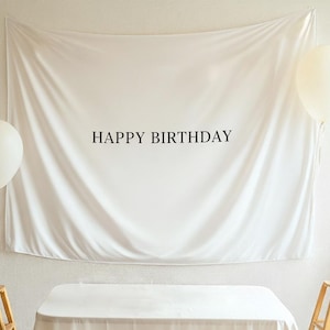 Może przedstawiać: Białe pł&oacute;tno z napisem "HAPPY BIRTHDAY" w czarnych literach. Widoczne są dwa białe balony, po jednym z każdej strony tła. Tło jest ułożone na stole.