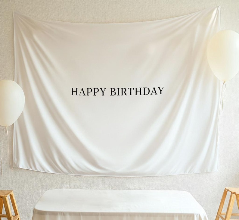 Może przedstawiać: Białe pł&oacute;tno z napisem "HAPPY BIRTHDAY" w czarnych literach. Widoczne są dwa białe balony, po jednym z każdej strony tła. Tło jest ułożone na stole.