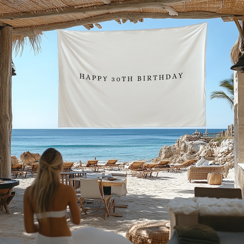 Puede incluir: Una pancarta blanca con el texto "HAPPY 30TH BIRTHDAY" en letras negras cuelga sobre un fondo de playa y oc&eacute;ano. La escena est&aacute; enmarcada por vigas de madera y un cielo azul claro. Se ven tumbonas y mesas.