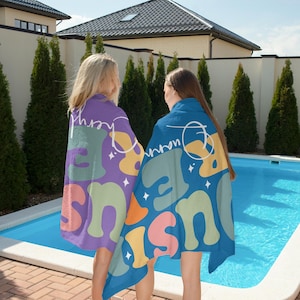 May include: Two people wearing colorful beach towels by a pool. The towels feature a retro design with the words "Just Be Here" in a playful font, along with abstract shapes in shades of purple, blue, green, orange, and yellow. The towels are perfect for a day at the beach or pool.
