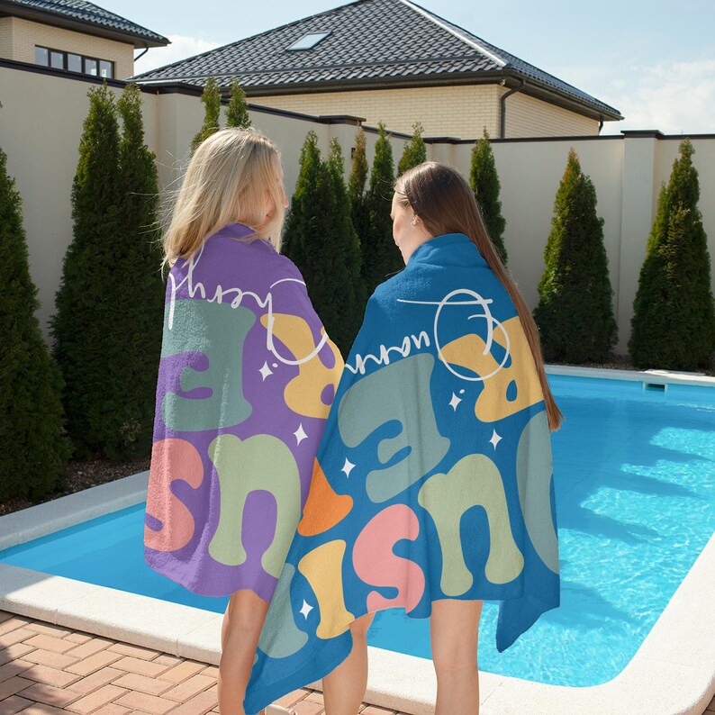 May include: Two people wearing colorful beach towels by a pool. The towels feature a retro design with the words "Just Be Here" in a playful font, along with abstract shapes in shades of purple, blue, green, orange, and yellow. The towels are perfect for a day at the beach or pool.