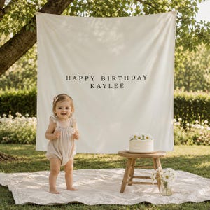 May include: A large, off-white fabric backdrop featuring the black printed text HAPPY BIRTHDAY KAYLEE. The banner is suspended outdoors, serving as a decorative display for a birthday celebration, positioned behind a small wooden stool and cake.