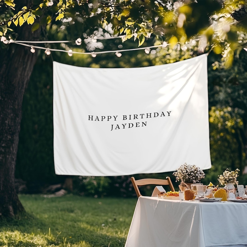 Puede incluir: Una pancarta blanca con el texto "HAPPY BIRTHAY JAYDEN" cuelga al aire libre. La pancarta est&aacute; colocada sobre una mesa preparada para una fiesta, con una silla de madera y arreglos florales. Luces de cadena est&aacute;n colgadas arriba.
