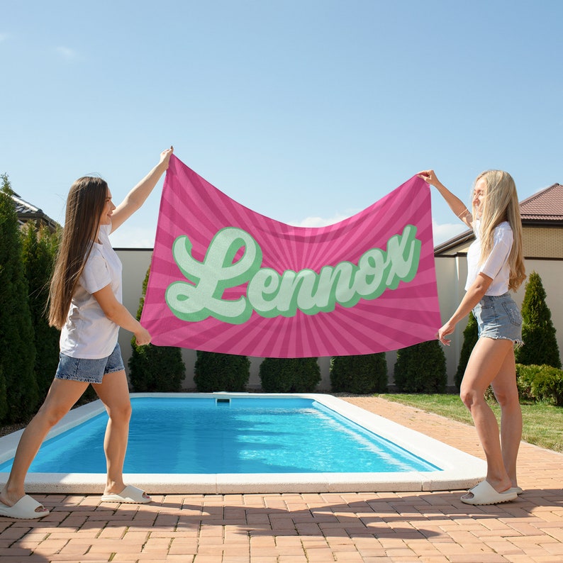 May include: A pink beach towel with the name "Lennox" in a retro font. The towel has a sunburst design and is held up by two people near a swimming pool on a sunny day. The towel is a vibrant accessory for the beach or pool.