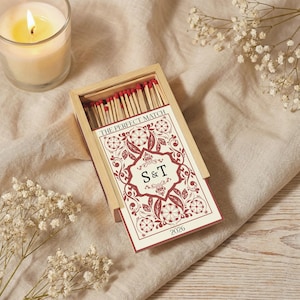 May include: A wooden matchbox with a decorative card featuring the text "THE PERFECT MATCH" and the initials "S&T". The card has a burgundy floral design and the number "2026". Red-tipped matches are visible inside the box. A lit candle and flowers are in the background.