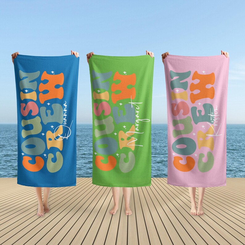 May include: Three beach towels with the text "COUSIN CREW" in orange, green, and blue, with names in white script. The towels are held up against a backdrop of the ocean and sky, with a wooden dock in the foreground.