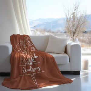 Peut inclure: Une couverture en polaire de couleur rouille avec un motif floral blanc et le nom "lindsay" imprim&eacute; dessus. La couverture est pos&eacute;e sur un canap&eacute; blanc avec un coussin blanc.