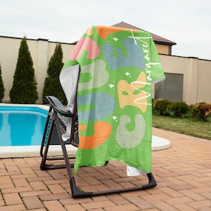 May include: A green beach towel draped over a chair, featuring a colorful abstract design with shapes in orange, blue, and pink. The towel has the name "Margaret" written in white script. The towel is on a chair near a pool.