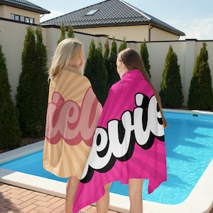 May include: Two beach towels with text. One towel is tan with a retro sunburst design and the word "ice" in pink. The other towel is pink with a striped pattern and the word "evie" in white with a black outline. The towels are near a swimming pool.