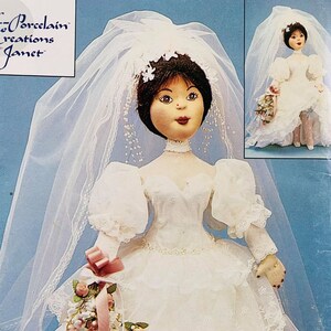 May include: A porcelain doll dressed in a white wedding gown with a veil and holding a bouquet of pink roses. The doll has black hair and brown eyes. The doll is wearing a white lace collar and a white lace trim on the sleeves of the dress. The doll is standing on a white background.