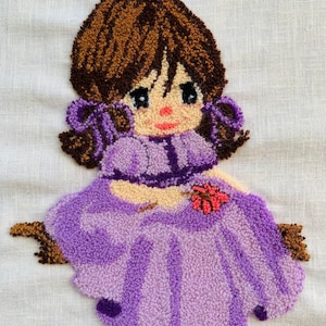 May include: A purple and brown yarn doll with a flower in her hand. The doll is wearing a purple dress and has brown hair.