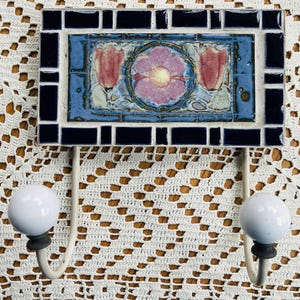 May include: A decorative wall hook with a rectangular ceramic tile featuring a floral design in pink, blue, and white. The tile is framed by dark blue mosaic tiles. Two white ceramic knobs with metal hooks are attached below.
