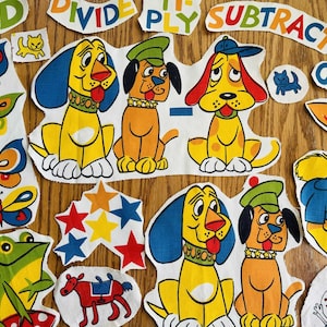 May include: A collection of colorful fabric cutouts featuring cartoon dogs, butterflies, stars, a frog, and a horse. The cutouts include words like ADD, DIVIDE, and SUBTRACT. The dogs are yellow and orange with blue and green accents.