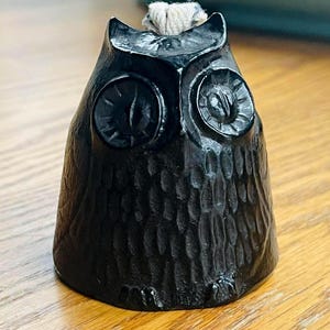 May include: A black owl-shaped candle holder with a textured surface. The owl has large, detailed eyes and a small wick protruding from the top. The candle holder is made of a dark material and sits on a wooden surface.