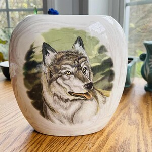 May include: A white ceramic vase with a detailed painting of a wolf's head. The wolf is depicted with gray and brown fur, yellow eyes, and its tongue slightly out. The background features green foliage.
