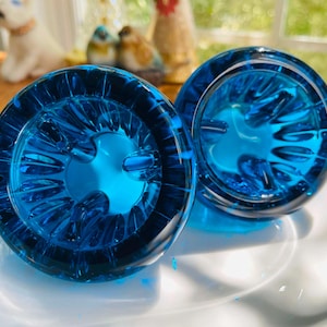 May include: Two circular, translucent blue glass objects with a textured, radial pattern on the underside. Each piece features a series of raised, petal-like ridges radiating from the center toward the outer rim, creating a deep, vibrant blue hue.