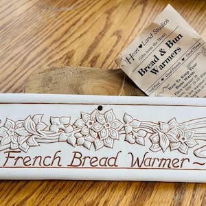 May include: Cream-colored ceramic French bread warmer with floral and swirl design. The words "French Bread Warmer" are inscribed in brown. A tag reads "Bread & Bun Warmers."