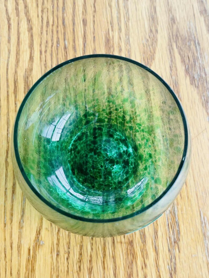 Puede incluir: Un cuenco redondo de vidrio con un dise&ntilde;o moteado verde y amarillo. El cuenco tiene un borde verde oscuro y un interior claro y reflectante. El cuenco est&aacute; sobre una superficie de madera.