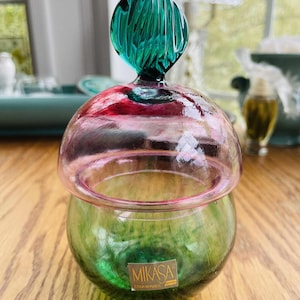 Puede incluir: Un tarro de cristal Mikasa Rep&uacute;blica Checa con base verde y tapa rosa y transparente. La tapa est&aacute; rematada con un adorno de cristal verde en forma de hoja. El tarro est&aacute; sobre una superficie de madera.