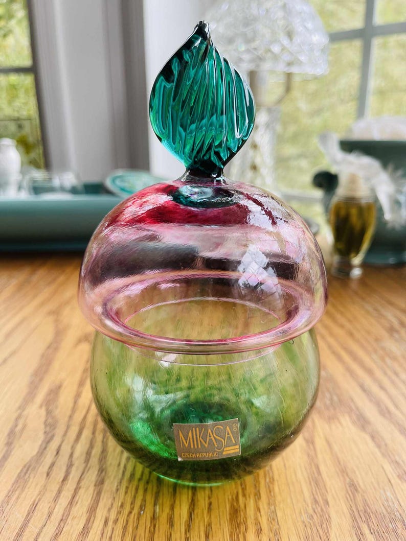 Puede incluir: Un tarro de cristal Mikasa Rep&uacute;blica Checa con base verde y tapa rosa y transparente. La tapa est&aacute; rematada con un adorno de cristal verde en forma de hoja. El tarro est&aacute; sobre una superficie de madera.