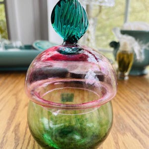 Puede incluir: Un tarro de cristal decorativo con base verde y tapa en forma de c&uacute;pula rosa y roja. La tapa est&aacute; rematada con un adorno de cristal texturizado de color verde azulado. El tarro est&aacute; sobre una superficie de madera.