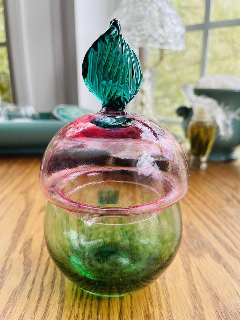 Puede incluir: Un tarro de cristal decorativo con base verde y tapa en forma de c&uacute;pula rosa y roja. La tapa est&aacute; rematada con un adorno de cristal texturizado de color verde azulado. El tarro est&aacute; sobre una superficie de madera.