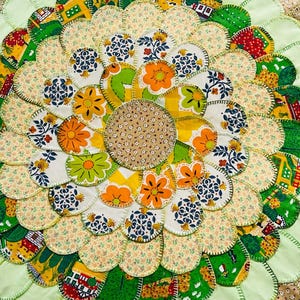 May include: A circular fabric mat featuring a layered petal design. The center is a beige floral print, surrounded by concentric rings of fabric petals in various patterns including orange flowers, green landscapes, blue floral motifs, and yellow prints.