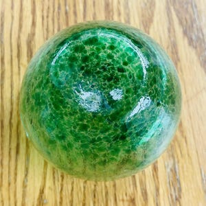 Puede incluir: Un objeto decorativo redondo de vidrio verde con una superficie texturizada. El objeto es de un tono verde vibrante y parece estar hecho de vidrio. El objeto est&aacute; sobre una superficie de madera.